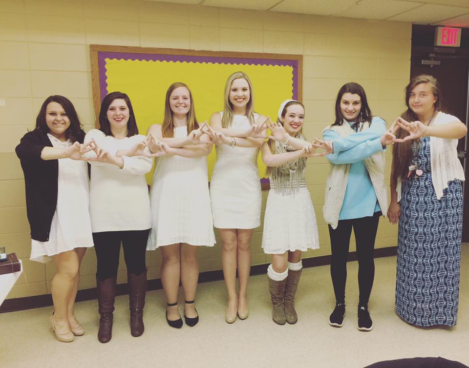 Members of Alpha Delta Chi dressed in white holding their hands together to form chapter hand sign.